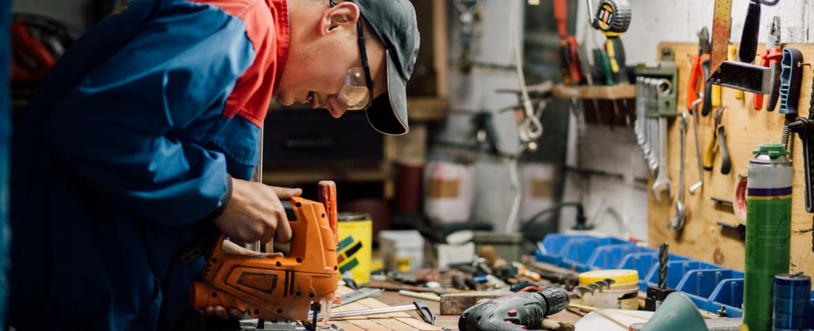 Ein Handwerker sägt in einer Werkstatt Holz mit einer Stichsäge. Er trägt Schutzbrille und Arbeitskleidung.