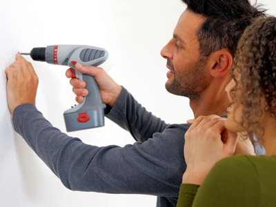 Ein Foto zeigt zwei Personen beim Anbringen einer Wandhalterung mit einer Akkubohrmaschine. Der Mann im Vordergrund fixiert eine Schraube in der Wand, während die Frau interessiert zusieht. Die Szene steht beispielhaft für die Montage eines Spiegels mit einem French-Cleat-System