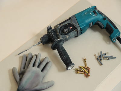 Auf einer Baustellenunterlage liegen eine Bohrmaschine, ein Paar Arbeitshandschuhe und sortierte Schrauben. Die Maschine ist gebraucht und mit einem Bohrer ausgestattet.