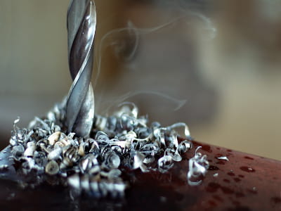 Ein Bohrer hat sich durch Metall gearbeitet und ist von heißen Metallspänen umgeben. Aus den Spänen steigt Dampf oder Rauch auf, was auf hohe Reibungshitze hinweist.