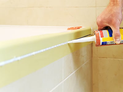 Foto zeigt eine Hand, die mit einer Kartuschenpistole weiße Silikonfuge entlang des Badewannenrands aufträgt, der Bereich ist mit Kreppband abgeklebt, um eine saubere Kante beim Abdichten zu erzielen