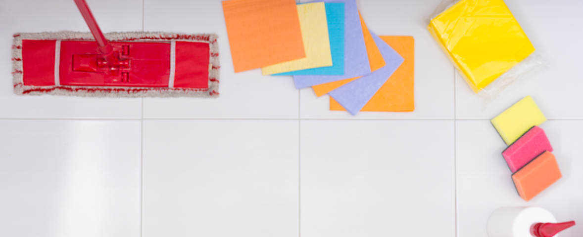 Eine Draufsicht zeigt die grundlegenden Reinigungsutensilien für die Badreinigung auf weißen Fliesen. Zu sehen sind ein Bodenwischer mit rotem Bezug, farbige Putzlappen, ein verpacktes Set gelber Tücher, drei Reinigungsschwämme in verschiedenen Farben und eine weiße Flasche mit roter Düse