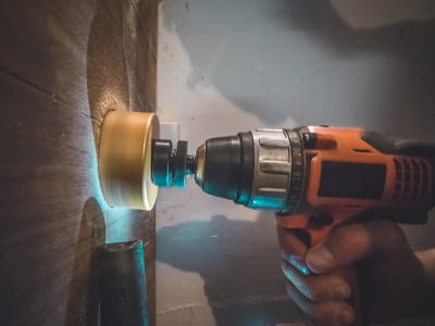 Eine Person bohrt mit einer orangefarbenen Akkubohrmaschine und einer Lochsäge in eine Wand. Die Szene ist schwach beleuchtet und zeigt eine präzise Arbeitssituation.