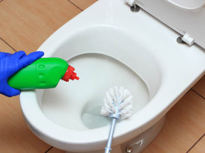 Eine Nahaufnahme zeigt die Reinigung einer Toilette. Eine Hand mit blauem Gummihandschuh trägt WC-Reiniger aus einer grünen Flasche mit roter Düse auf den Rand der Toilettenschüssel auf. Gleichzeitig wird mit einer weißen Toilettenbürste das Innere der Schüssel gereinigt