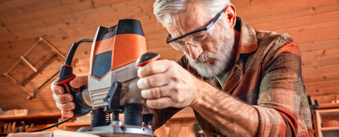 Ein älterer Mann mit Schutzbrille arbeitet konzentriert mit einer Oberfräse an einem Holzstück. Er befindet sich in einer warm beleuchteten Werkstatt mit Holzverkleidung.