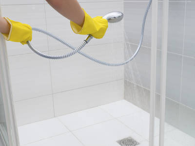 Eine Person mit gelben Gummihandschuhen hält eine Handbrause und spült die geflieste Duschkabine aus. Wasser läuft über den sauberen weißen Fliesenboden in den Ablauf, die Glaswände der Dusche sind geöffnet