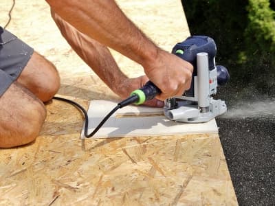 Eine Person arbeitet im Freien mit einer Oberfräse an einer OSB-Platte. Dabei entsteht feiner Holzstaub, der zur Seite wegfliegt.