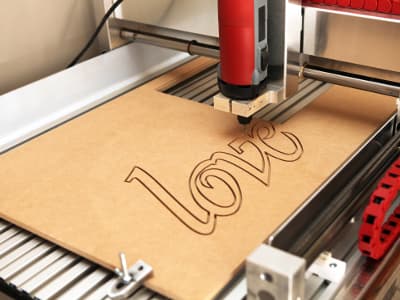 Eine CNC-Fräse graviert das Wort „love“ in eine Holzplatte. Der Fräskopf fährt präzise die Buchstabenformen entlang.