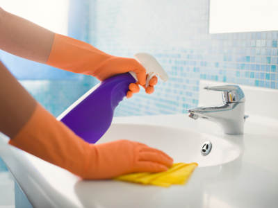 Eine Nahaufnahme zeigt die Reinigung eines Waschbeckens. Eine Hand mit orangenen Gummihandschuh trägt einen Reiniger aus einer lilafarbenen Flasche. Gleichzeitig wird mit einem gelben Lappen das Waschbecken gereinigt