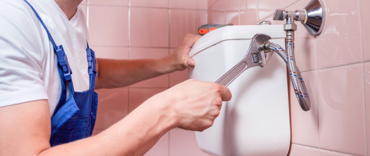 Ein Mann schließt einen flexiblen Zulaufschlauch mit einem verstellbaren Schraubenschlüssel an den Spülkasten eines Stand-WCs an. Der Spülkasten ist an rosa gefliesten Wänden montiert, auf dem Spülkasten liegt ein Schraubendreher