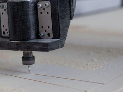 Eine CNC-Fräse bearbeitet eine helle Holz- oder Kunststoffplatte. Der Fräskopf schneidet präzise Linien in das Material, während feiner Staub entsteht.