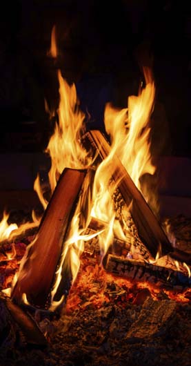 Nahaufnahme von lodernden Flammen in einem Kaminofen. Drei Holzscheite brennen in einer V-Form über glühender Asche. Die gelb-orange Flamme ist klar zu erkennen, das Holz ist bereits angesengt