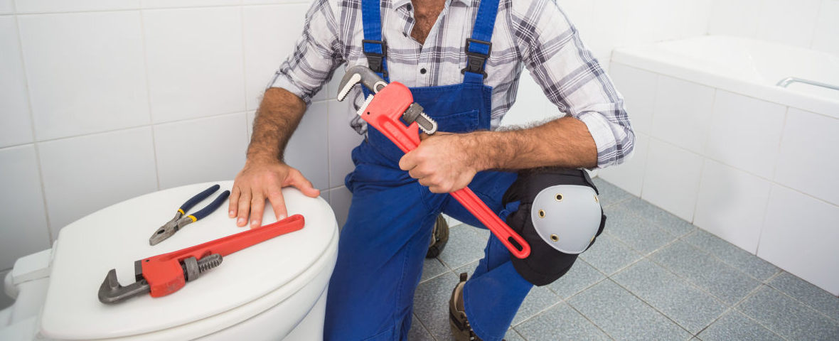 toilettendeckel-befestigen-header.jpg Ein Handwerker in blauer Latzhose befestigt einen Toilettendeckel. Der Mann kniet vor einem WC, auf dem ein roter und ein blauer Schraubenschlüssel liegen, in der Hand hält er eine Rohrzange. Der Raum ist vollständig gefliest, im Hintergrund ist eine Badewanne zu sehen