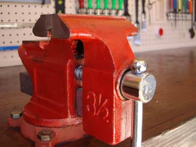 Ein massiver roter Schraubstock ist auf einer Werkbank montiert. Im Hintergrund sind weitere Werkzeuge ordentlich an einer Lochwand befestigt