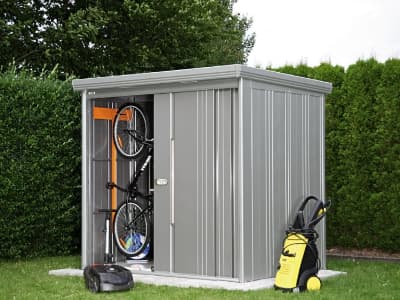Ein kompaktes Gerätehaus aus Metall mit Schiebetür steht auf einer Betonplatte im Garten. Darin befinden sich unter anderem zwei Fahrräder, davor ein Hochdruckreiniger und ein Mähroboter
