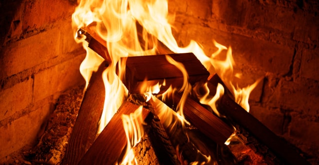 Offenes Feuer mit hellen Flammen und glühendem Holz in einem gemauerten Kamin. Die Holzscheite sind pyramidenförmig gestapelt, die Flammen schlagen gleichmäßig nach oben, die Steine im Hintergrund sind rötlich angestrahlt