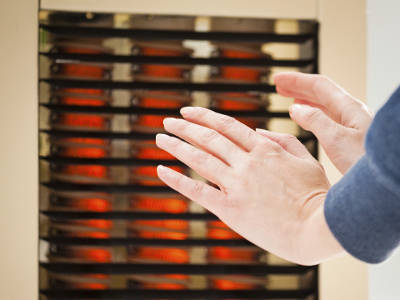 Zwei Hände werden vor einem eingeschalteten Heizlüfter mit rot glühenden Heizelementen gewärmt. Die Wärmequelle steht in einem Innenraum mit heller Wand