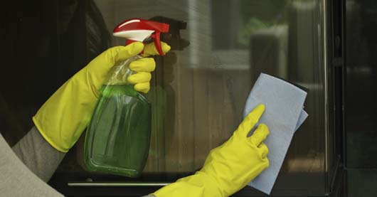 Reinigung einer Kaminofenscheibe mit gelben Gummihandschuhen, einer Sprühflasche mit grünem Reinigungsmittel und einem blauen Tuch. Die Glasoberfläche spiegelt das Licht leicht