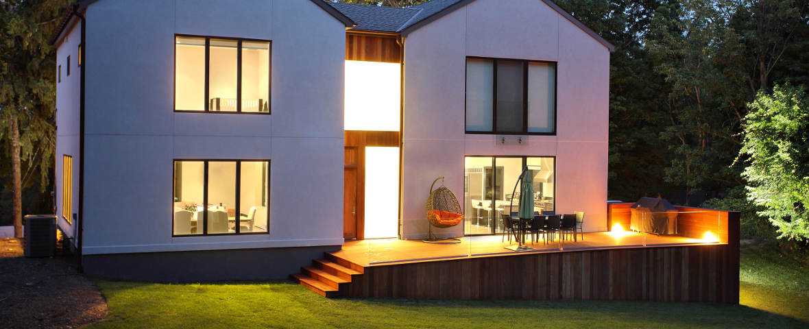 Eine moderne Terrasse mit Holzverkleidung ist dezent von integrierten Bodenleuchten beleuchtet. Das Haus im Hintergrund ist ebenfalls erleuchtet, auf der Terrasse stehen Gartenmöbel und ein Hängesessel