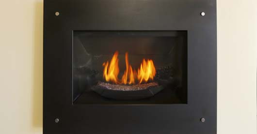 Moderner wandintegrierter Kaminofen mit schwarzem Metallrahmen und Sichtfenster. Hinter der Scheibe lodern gleichmäßige Flammen auf glühendem Brennmaterial. Die Wandfarbe ist hell und kontrastiert mit dem schwarzen Rahmen