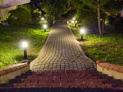 Aus der Egoperspektive blickt man am Treppenkopf diese hinunter und sieht einen steinernen Weg in einem Garten, der mit Lampen, die aus dem Boden ragen, beleuchtet ist