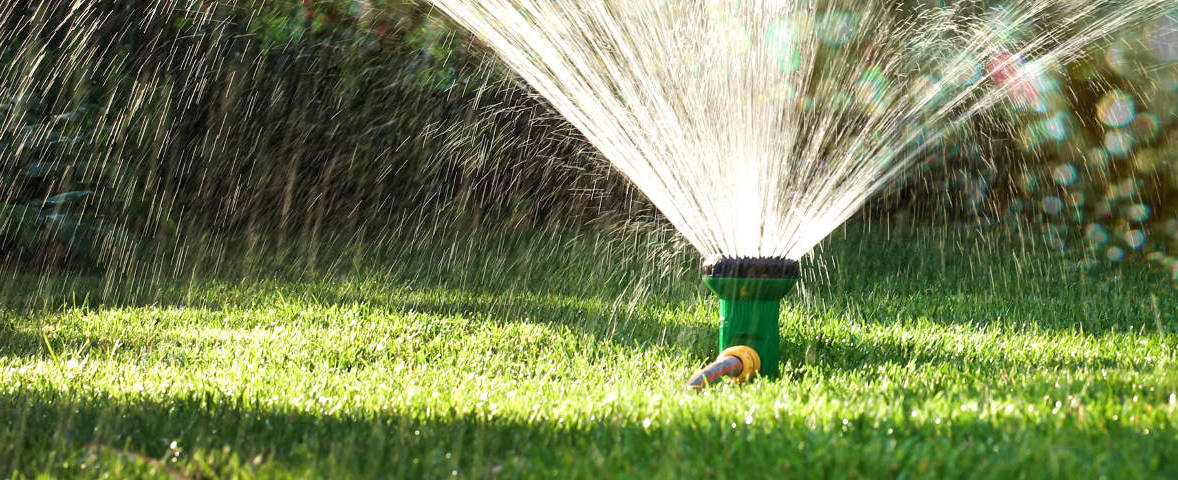 Nahaufnahme eines Rasensprengers, der an einem Wasserschlauch angeschlossen ist und fächerförmig ein Stück Rasen bewässert