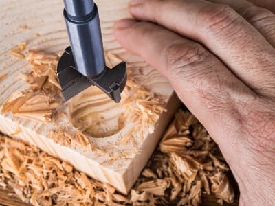 Ein Forstnerbohrer fräst ein rundes Loch in ein Holzstück, Späne liegen ringsum.