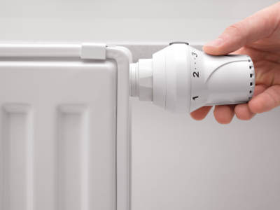 Eine Hand dreht an einem weißen Heizkörper-Thermostat, das auf Stufe 2 bis 3 eingestellt ist. Der Heizkörper ist ebenfalls weiß und steht vor einer hellen Wand. Die Szene vermittelt den Hinweis auf richtiges Heizverhalten zur Vorbeugung von Schimmel