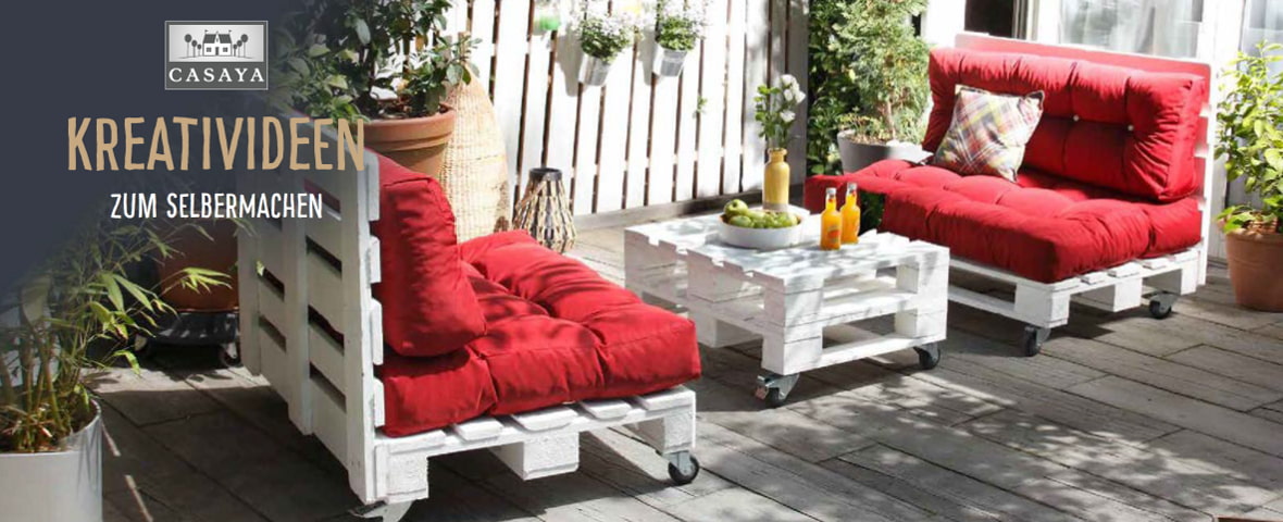 Eine gemütliche Terrassenszene zeigt selbst gebaute Palettenmöbel bestehend aus zwei Sesseln und einem Couchtisch mit Rollen. Die Paletten sind weiß gestrichen und mit dicken roten Polstern ausgestattet. Auf dem Tisch stehen Getränke und Obst, im Hintergrund stehen Pflanzen und Holzwände. Links ist das Casaya-Logo. Darunter steht: Kreativideen zum Selbermachen