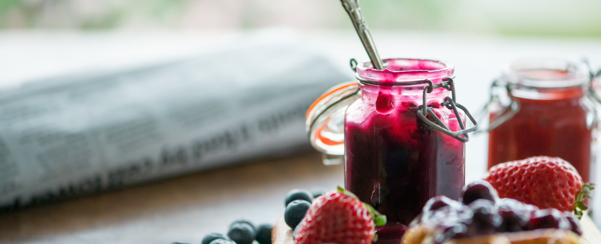 Offenes Glas mit dunkler Beerenmarmelade und Löffel, umgeben von frischen Erdbeeren, Heidelbeeren und einem Gebäckstück mit Fruchtbelag