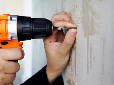 Eine Person schraubt mit einem orangefarbenen Akkuschrauber eine Schraube in eine Wand. Die Schraube wird mit der freien Hand gehalten und ausgerichtet.