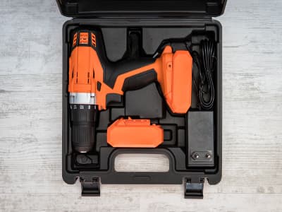Ein orangefarbener Akkuschrauber liegt in einem schwarzen Transportkoffer. Neben dem Gerät befinden sich ein Ladegerät und ein Ersatzakku.