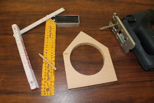 Auf einer Holzarbeitsfläche liegen Werkzeuge und vorbereitete Bauteile für ein Vogelhaus. Zu sehen sind ein zugesägtes Vorderteil mit rundem Ausschnitt, ein Zollstock, ein Winkel, ein Bleistift und eine Stichsäge. Die Werkzeuge dienen zum Anzeichnen und exakten Sägen der Holzelemente