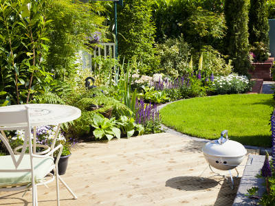 Eine helle Holzterrasse mit weißem Metalltisch und Stuhl geht in einen sorgfältig gepflegten Garten mit runder Rasenfläche und üppigen Blumenbeeten über. Ein kleiner Kugelgrill steht auf der Terrasse