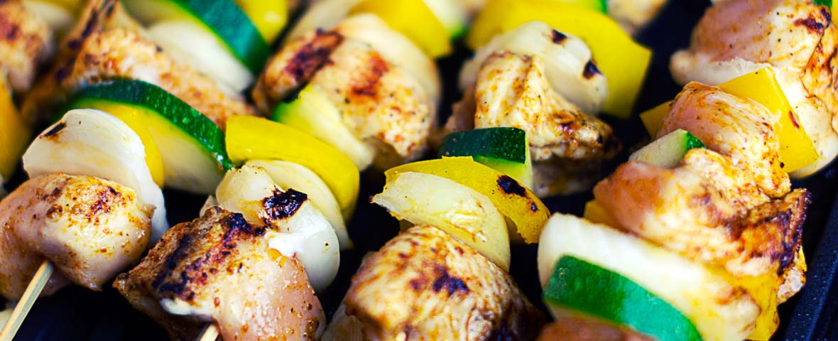 Mehrere bunte Grillspieße mit gewürzten Hähnchenstücken, Zucchini, Paprika und Zwiebeln liegen nebeneinander auf einem Grill und sind bereits leicht gebräunt