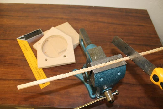 Auf einer Werkbank wird eine dünne Holzrundstange in einem Schraubstock eingespannt und mit einer Handsäge zugeschnitten. Im Hintergrund liegen vorbereitete Holzbauteile für ein Vogelhaus mit rundem Ausschnitt, ein Winkel und ein Zollstock