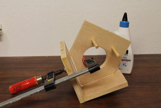 Auf einer Werkbank werden die beiden Dachhälften eines Vogelhauses mit einer Schraubzwinge fixiert und verleimt. Die Vorderseite mit rundem Ausschnitt und eingesetzten Sitzstangen ist bereits montiert. Im Hintergrund steht eine Flasche Holzleim