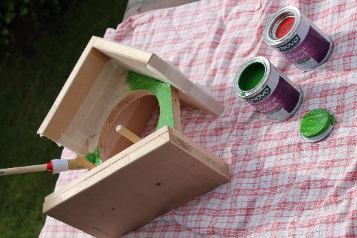 Auf einem karierten Tuch liegt ein teilweise montiertes Vogelhaus, das mit grüner Holzfarbe gestrichen wird. Ein Pinsel ist in die frische Farbe eingetaucht und liegt am Rand der Frontplatte mit rundem Ausschnitt. Zwei geöffnete Farbdosen in Grün und Rot stehen daneben bereit