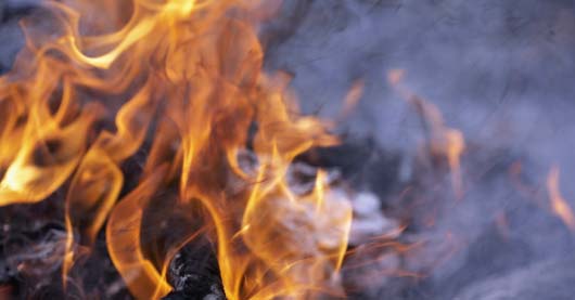 Nahaufnahme von flackernden Flammen mit orange-gelben Farben vor dunklem, rauchigem Hintergrund. Einzelne Rauchschwaden ziehen durch das Bild, die Glut ist nur angedeutet sichtbar