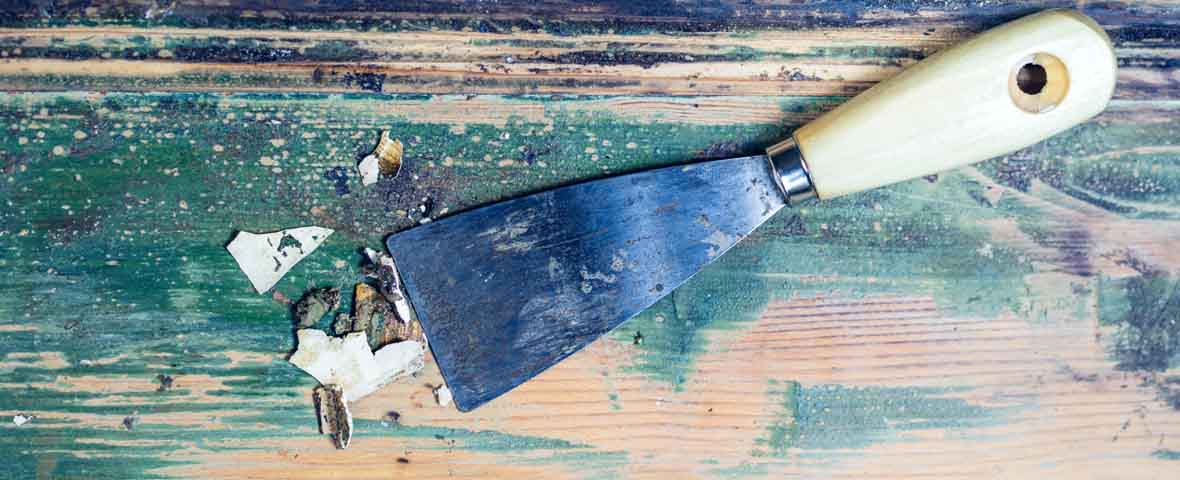 Foto eines Untergrunds aus Holz mit abgeblätterter Farbe, darauf liegt ein Spachtel und es sind bereits Lack- und Farbreste zu sehen, die entfernt wurden
