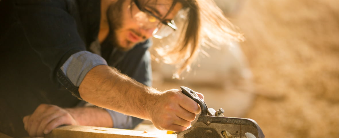 Ein Mann mit Schutzbrille arbeitet konzentriert mit einem Hobel an einem Holzstück.