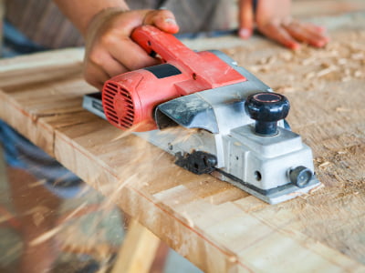Eine Person bearbeitet eine Holzoberfläche mit einem elektrischen Handhobel. Holzspäne fliegen während des Arbeitens zur Seite.