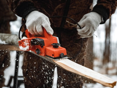 Eine Person mit Handschuhen bearbeitet im Freien ein Holzbrett mit einem elektrischen Hobel. Holzspäne fliegen durch die winterliche Umgebung.