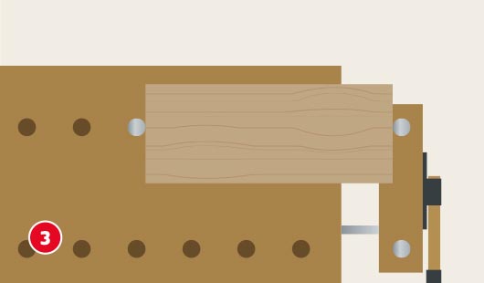 Eine schematische Darstellung zeigt ein rechteckiges Holzstück, das von oben betrachtet mittig auf einer Werkbank eingespannt ist. Zwei runde Spannbolzen und eine Schraubzwinge fixieren das Werkstück sicher. Die Oberfläche der Werkbank ist mit mehreren Bohrungen versehen. Unten links ist ein roter Kreis mit einer weißen 3