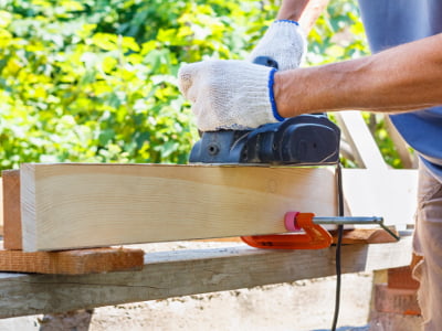 Eine Person glättet mit einem Bandschleifer ein eingespanntes Holzbrett im Freien. Das Holz ist auf einer Werkbank fixiert, und die Person trägt Arbeitshandschuhe.