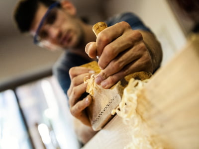 Ein Mann mit Schutzbrille bearbeitet konzentriert ein Holzstück mit einem Handhobel. Dabei entstehen feine, aufgerollte Holzspäne.