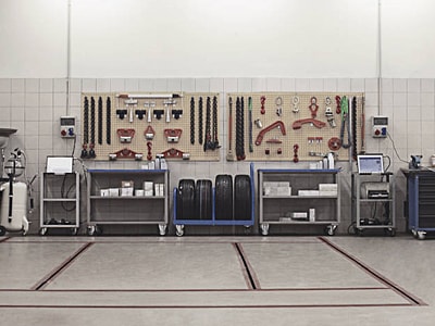 Eine modern ausgestattete Werkstatt zeigt Werkzeugwände mit geordnetem Equipment, mehrere Werkbänke auf Rollen sowie Computerarbeitsplätze. In der Mitte befinden sich Reifen und Kisten mit Material.