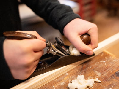 Zwei Hände führen einen Handhobel über ein Holzstück. Dabei lösen sich feine, helle Holzspäne von der Oberfläche.