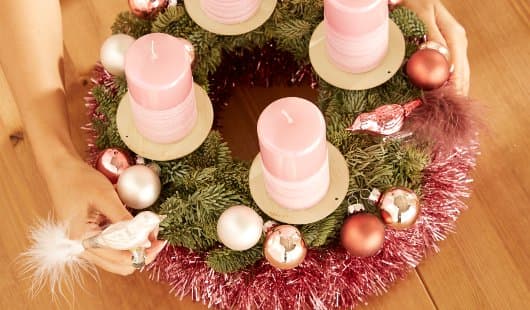 Ein Adventskranz mit rosafarbenen Kerzen und Tannenzweigen wird mit glänzenden Weihnachtskugeln in Rosa, Weiß und Kupfer sowie kleinen Vogelclips und flauschigen Elementen geschmückt. Zwei Hände platzieren die Dekoration rund um die Kerzen auf dem Kranz, der auf einem Holztisch liegt