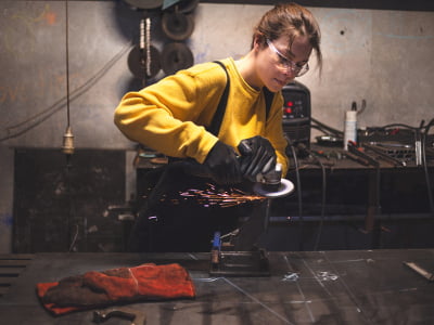 Eine Frau schleift in einer Werkstatt Metall mit einem Winkelschleifer. Funken fliegen